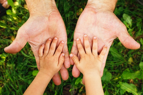 Close-up of a child’s small hands resting on top of an adult’s open palms against a background of green grass.