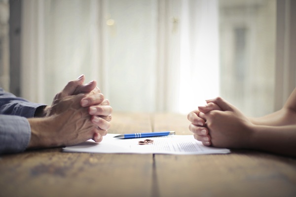 Two people sit across from each other with hands clasped over divorce papers and wedding rings on a table.