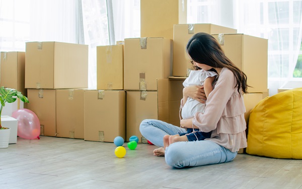 Mother holding her young child while sitting on the floor surrounded by moving boxes.