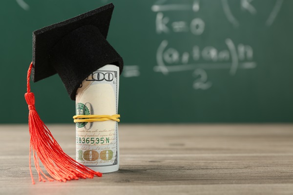 Rolled-up hundred-dollar bill wearing a graduation cap with a red tassel, symbolizing student loans or college tuition costs, placed in front of a chalkboard with math equations.
