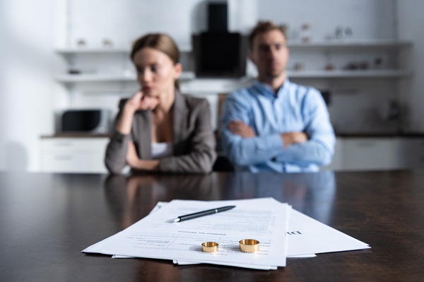 A pair of gold wedding rings and a pen rest on a "Petition for Divorce" document, with a blurred, frustrated couple sitting in the background.